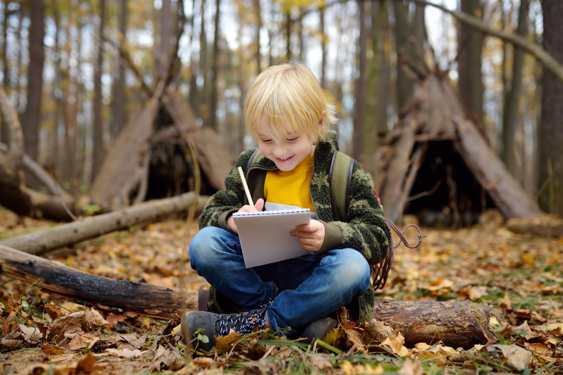 A child sitting on a log in the woods, writing in a notepad with a pencil.